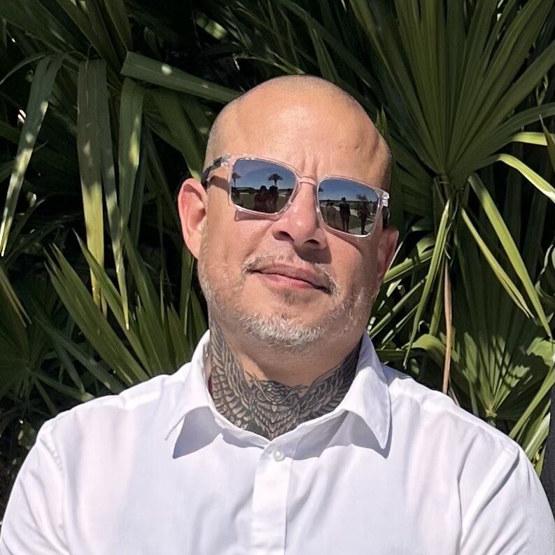 Gregory G. Camacho wearing sunglasses and a white shirt in front of tropical palm leaves